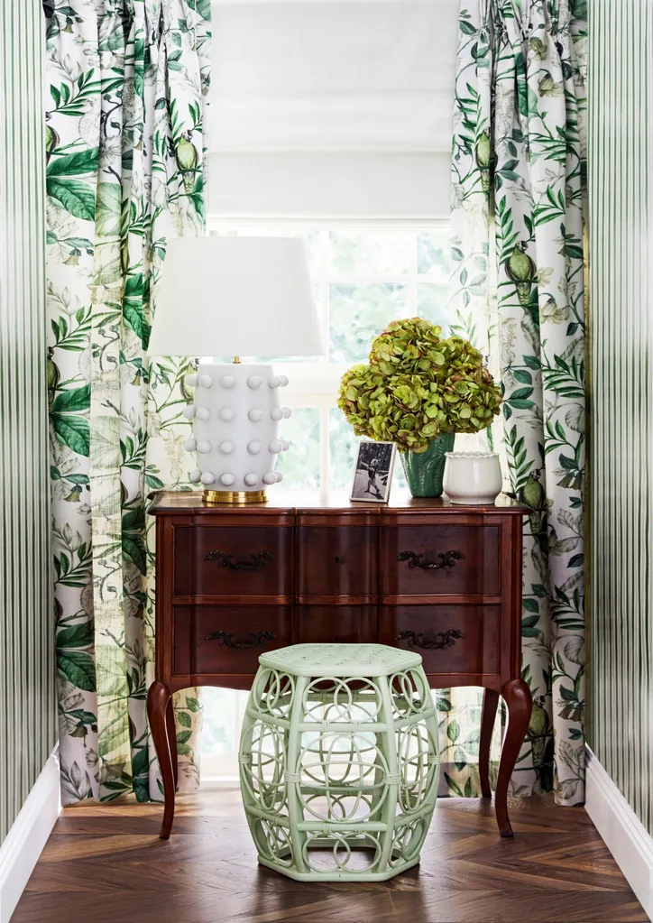 A window at the end of a hallway has green and white curtains with a botanical pattern over white blinds. A dark timber console table has a white table lamp on it, a vase of greenery and a green stool in front of it.