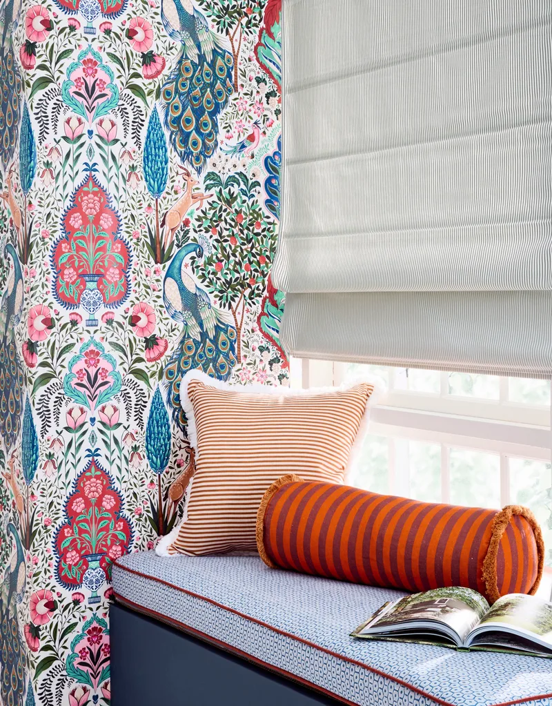 A window seat with cushions on top, a bright wallpaper covering the wall and a light grey blind on the window.