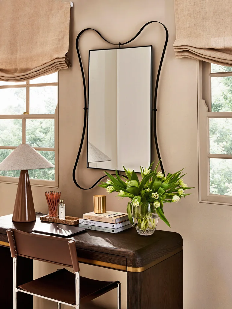 A light brown wall has a mirror with a black frame, with a timber desk in front. A vase of tulips are on the desk, beside coffee table books, a glass of freshly sharpened pencils and a lamp. Blinds in a fabric that is a similar shade to the walls are covering the top half of two windows.