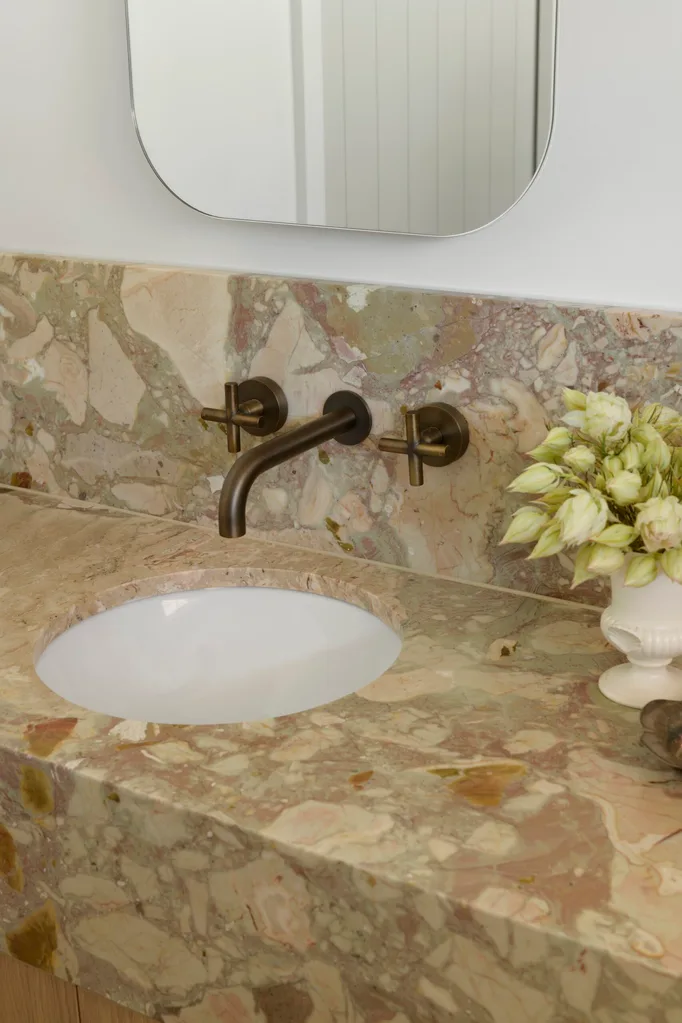 A close up of a bathroom vanity, with marble as the benchtop and splashback. The vanity has a deep apron, made from the same marble, which has warm tones complemented by antique brass tapware. A mirror is above the vanity, against a white wall, with a small vase of flowers to the side of the circular undermount basin on the vanity. The vanity is bold and a clear sign this bathroom was designed for personal preferences, not resale value.