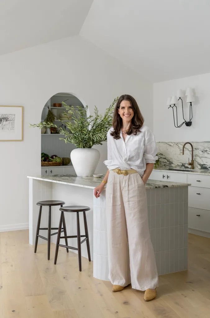 A kitchen with clear Spanish style influences, such as an archway leading from the kitchen and into the pantry, a vaulted ceiling and marble with bold veining on the benchtop and splashbacks. A woman with medium length brown hair stands in front of the kitchen island, smiling. She is the designer of the kitchen and aimed to please her clients, not design with resale value in mind.