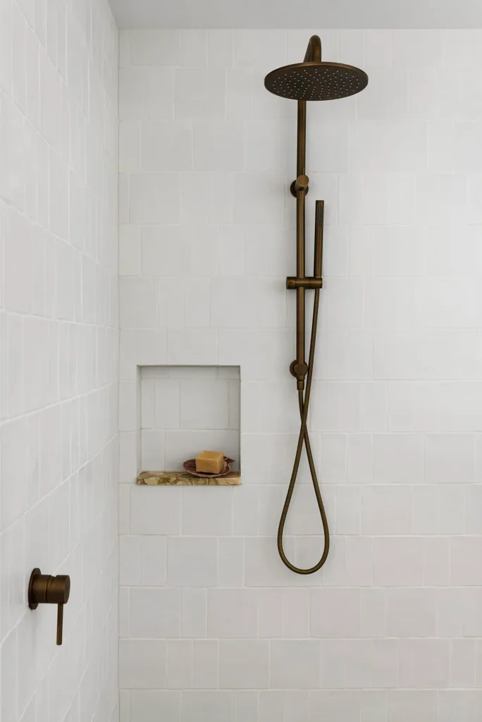 A close up of a shower with white square and rectangular tiles on the walls, a small square shower niche with a piece of marble comprising the bottom. Antique brass hardware makes up the shower head.
