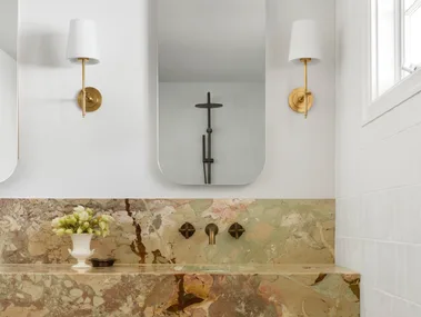 A bathroom vanity, with marble as the benchtop and splashback. The vanity has a deep apron, made from the same marble, which has warm tones complemented by antique brass tapware. Underneath the apron, oak joinery creates storage. Twin mirrors are positioned above twin basins in the vanity, with a wall sconce on either side.