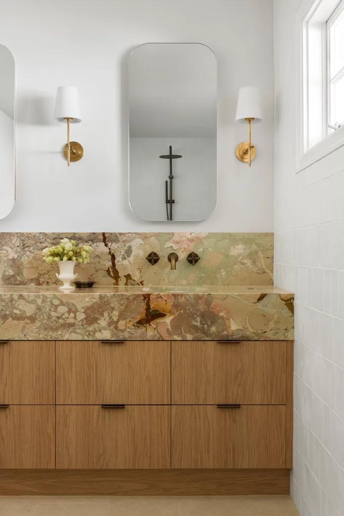 A bathroom vanity, with marble as the benchtop and splashback. The vanity has a deep apron, made from the same marble, which has warm tones complemented by antique brass tapware. Underneath the apron, oak joinery creates storage. Twin mirrors are positioned above twin basins in the vanity, with a wall sconce on either side.