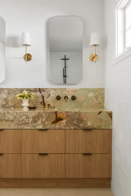 A bathroom vanity, with marble as the benchtop and splashback. The vanity has a deep apron, made from the same marble, which has warm tones complemented by antique brass tapware. Underneath the apron, oak joinery creates storage. Twin mirrors are positioned above twin basins in the vanity, with a wall sconce on either side.