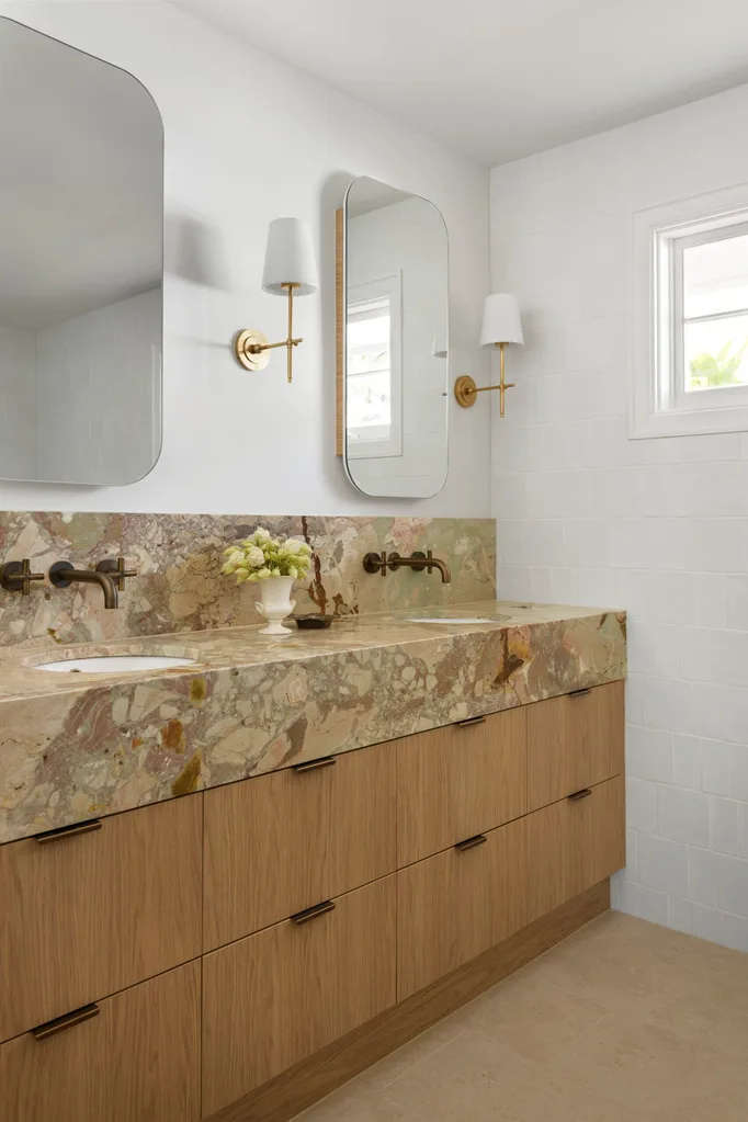 A bathroom vanity, with marble as the benchtop and splashback. The vanity has a deep apron, made from the same marble, which has warm tones complemented by antique brass tapware. Underneath the apron, oak joinery creates storage. Twin mirrors are positioned above twin basins in the vanity, with a wall sconce on either side. The vanity is bold and a clear sign this bathroom was designed for personal preferences, not resale value.