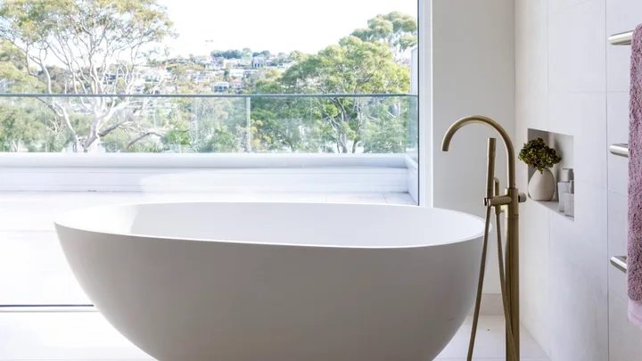 Large pavers cover the floor and wall in this ensuite bathroom, where a large oval-shaped freestanding bath takes centre stage in front of a large window with bushland views.