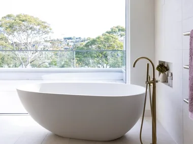 Large pavers cover the floor and wall in this ensuite bathroom, where a large oval-shaped freestanding bath takes centre stage in front of a large window with bushland views.