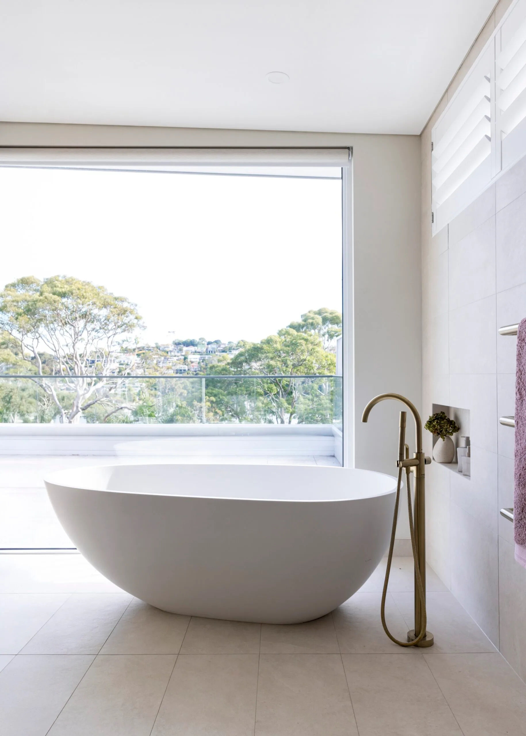 Large pavers cover the floor and wall in this ensuite bathroom, where a large oval-shaped freestanding bath takes centre stage in front of a large window with bushland views.