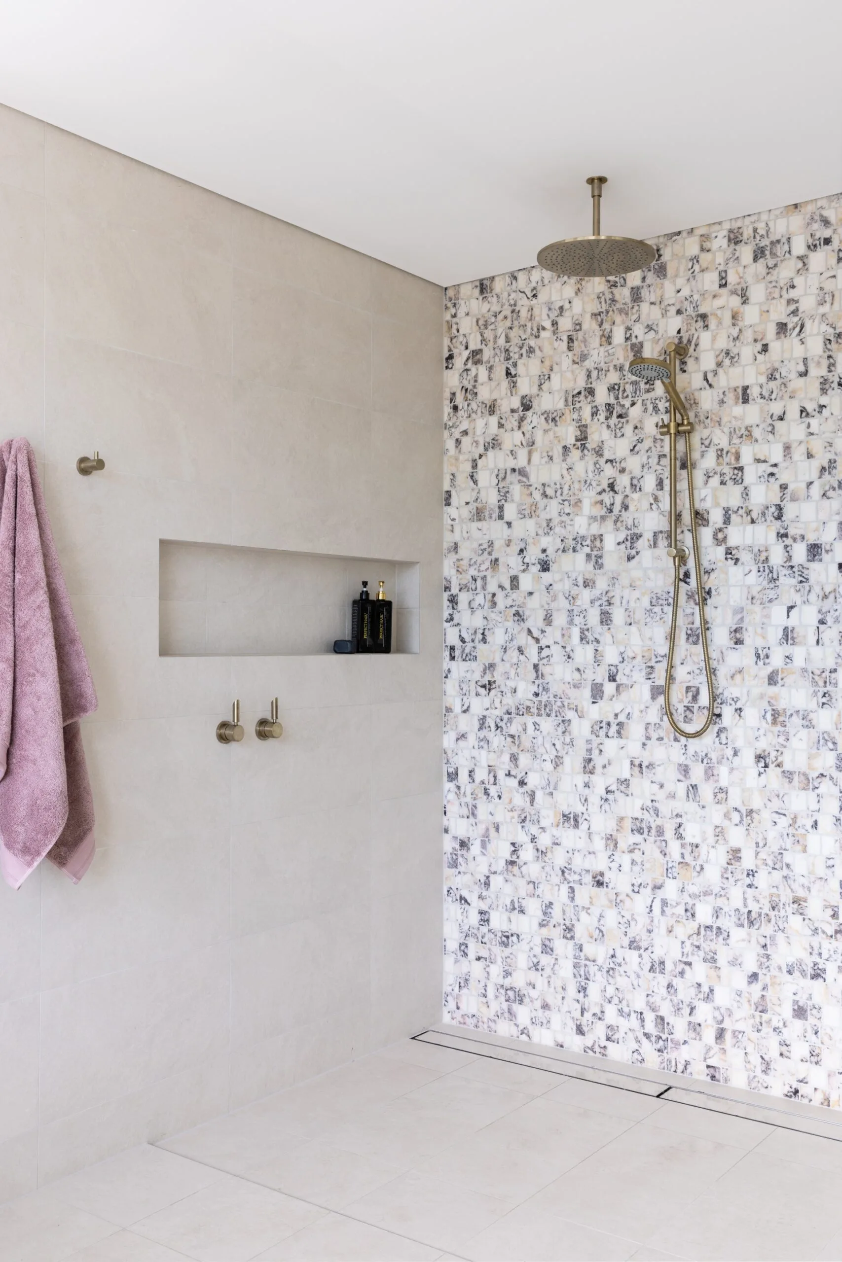 A large shower area with warm-toned pavers, that are large, covering the floor and one wall (including the shower niche). Brushed brass tapware is in the shower and there is a feature wall of honed marble mosaic tiles which have a soft pink hue, mixed with warm greys.