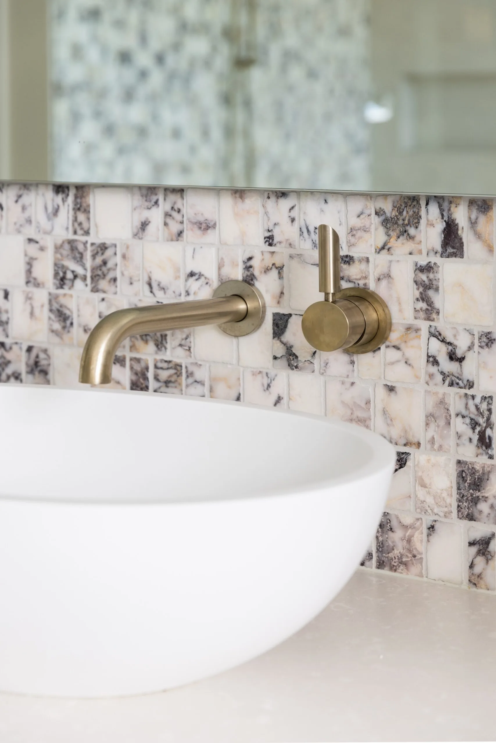 A close up photo of an above-counter basin, which is white with an oval shape. Brushed brass tapware is fitted into the wall above the basin, which is clad in honed marble mosaic tiles which have a soft pink hue, mixed with warm greys. Above this is a frameless mirror.