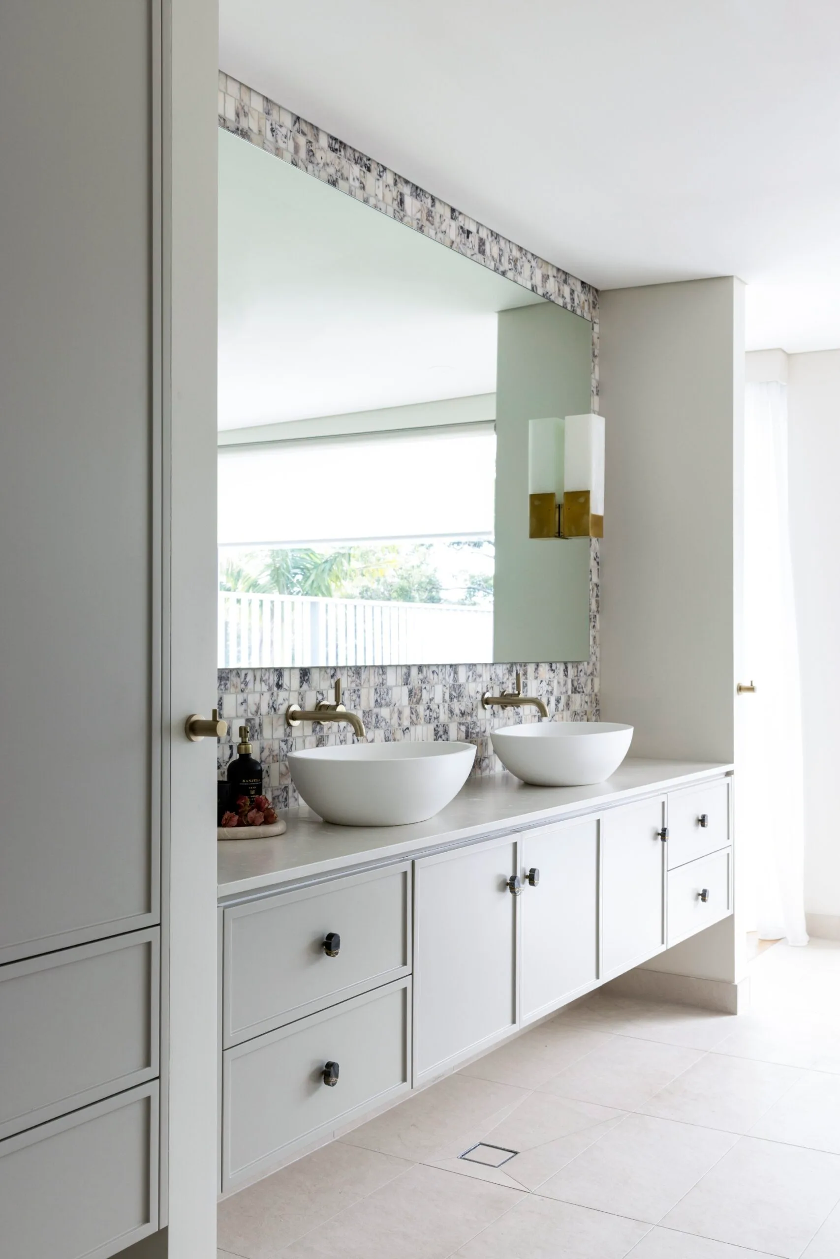 Joinery in an ensuite bathroom, which has a shaker profile, with a white benchtop and two white, oval-shaped above-counter basins on top. A large mirror spans the length of the wall above the joinery. Brushed brass tapware is fitted into the wall above the basins. The wall is clad in honed marble mosaic tiles which have a soft pink hue, mixed with warm greys. Large neutral tiles cover the floor.