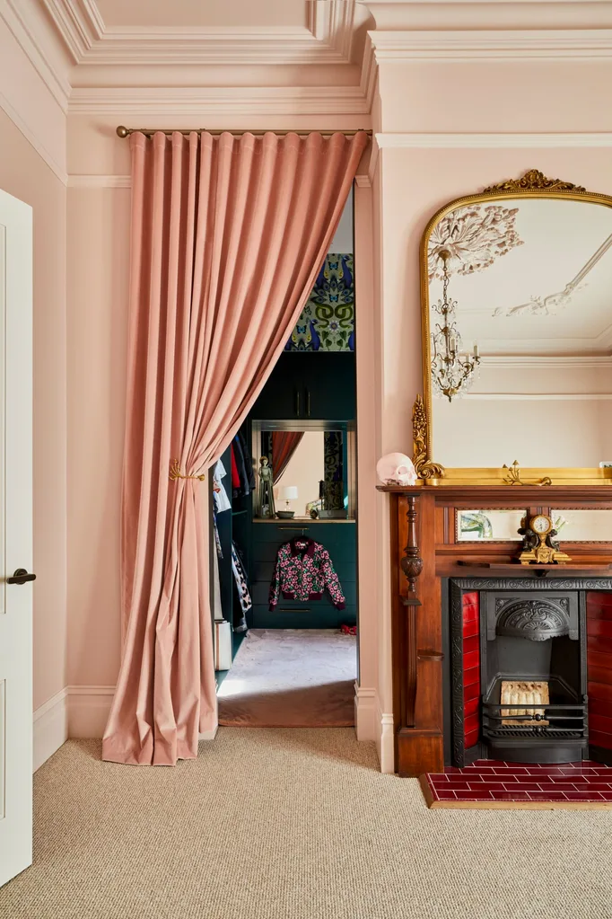 When Fleur Harris worked on her home renovation she painted her main bedroom pink and used a pink curtain to divide the space between her bedroom and walk-in robe. A fireplace has an ornate gold mirror positioned above.
