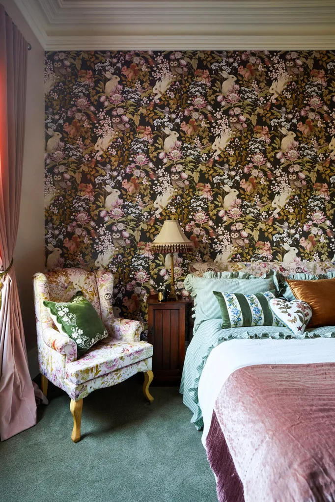 The guest bedroom in Fleur Harris' house. A feature wall has wallpaper with a green, pink and green pattern. The pattern is floral with woodland creatures such as rabbits, squirrels and owls. The bedlinen is pink and blue, with a wingback armchair in a pink floral pattern beside.