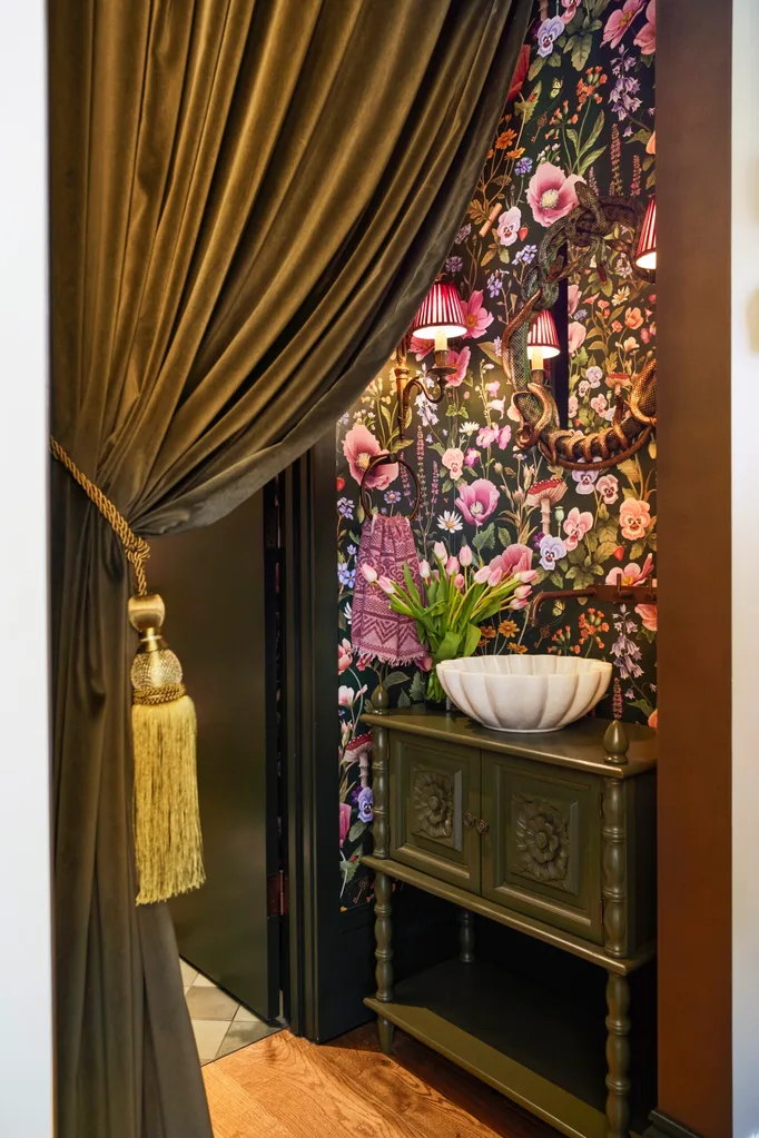 The powder room in Fleur Harris' home is separated by a large, green curtain. Green joinery is positioned beneath a white sink, while pink and green floral wallpaper covers the walls.