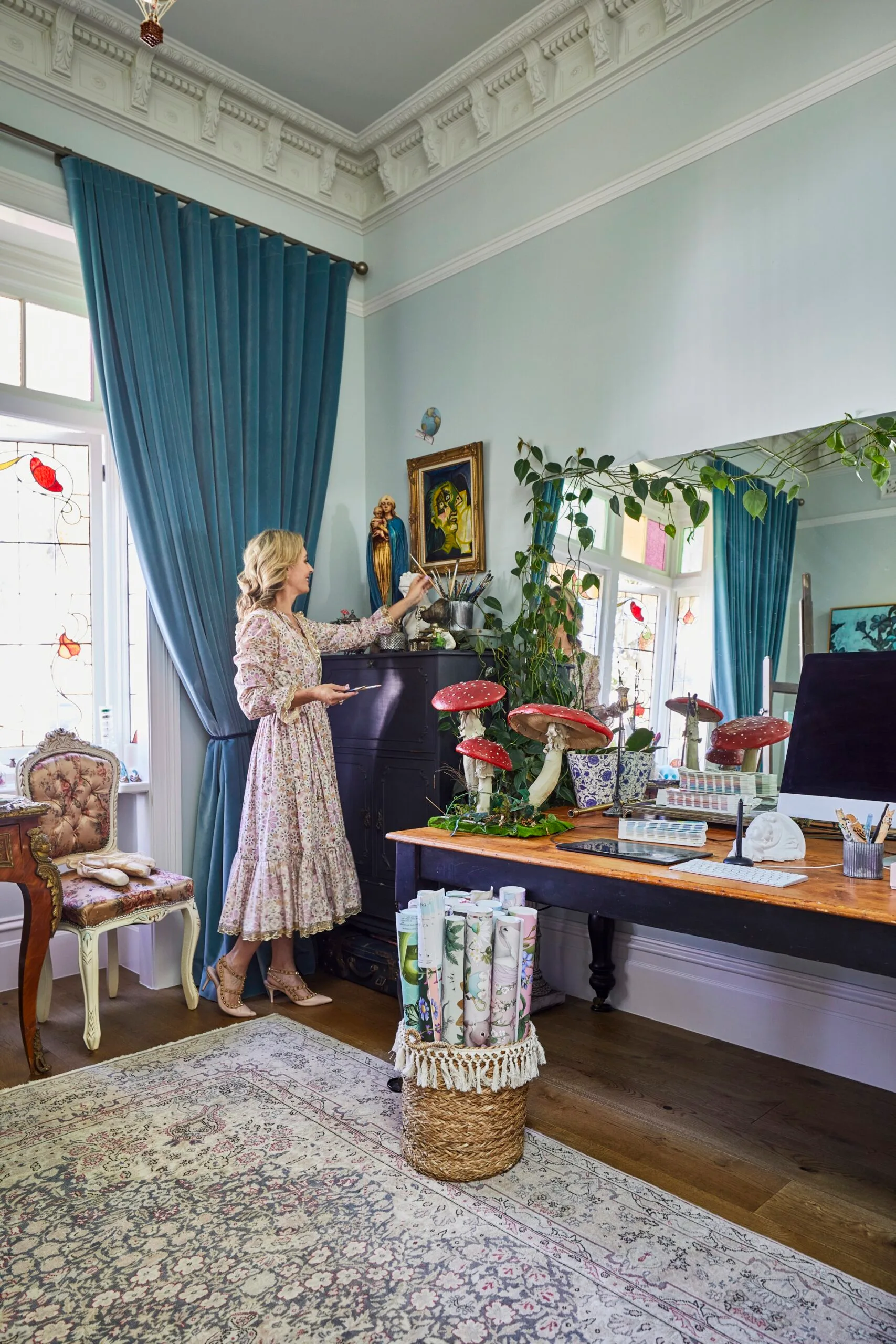 A blonde woman, Fleur Harris, in her home studio which has a large rug on the floor, blue walls and curtains, a stained glass window and lots of art decorating the space. During her home renovation, Fleur decorated with antiques and even props from photoshoots, including the fairytale mushroom statues on her desk.