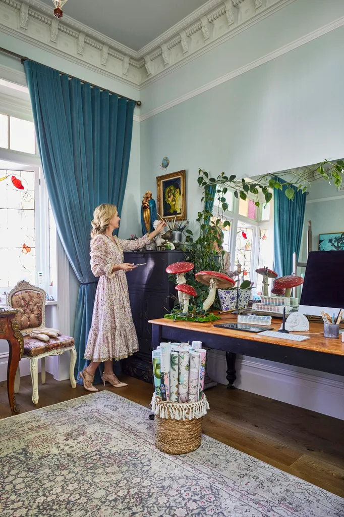 A blonde woman, Fleur Harris, in her home studio which has a large rug on the floor, blue walls and curtains, a stained glass window and lots of art decorating the space. During her home renovation, Fleur decorated with antiques and even props from photoshoots, including the fairytale mushroom statues on her desk.