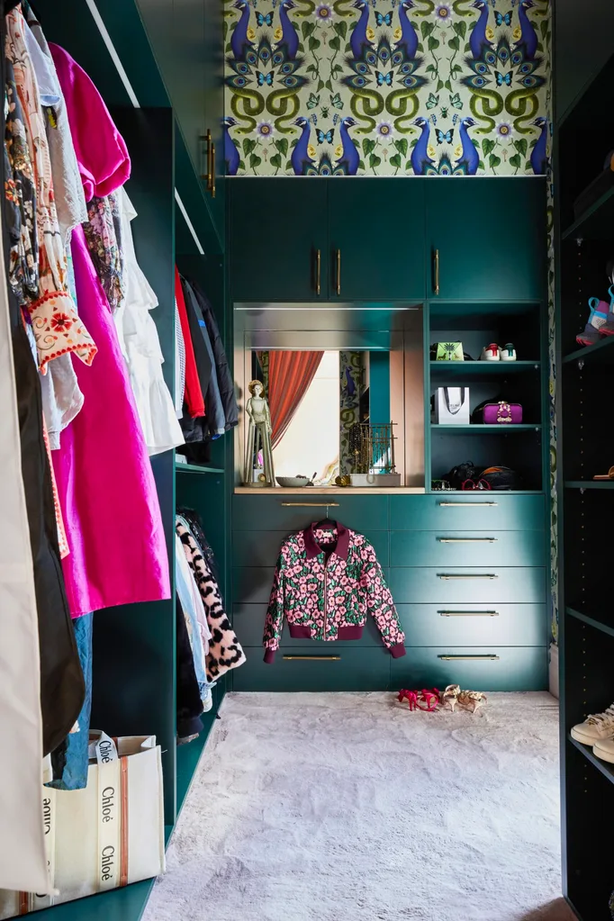 Fleur Harris' walk-in-robe, with a deep teal joinery, brass handles and wallpaper that is patterned with blue and green snakes, peacocks and butterflies. Her clothes, which are hanging up on the left side of the walk-in-robe, are colourful and mostly pink.