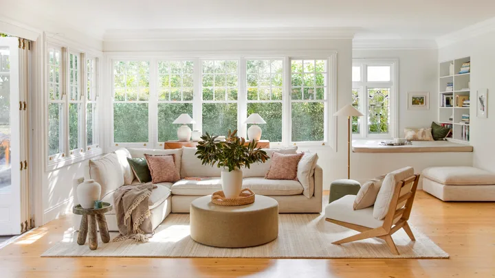 A living room with light timber floorboards, white walls and large windows that look out on to greenery. A cream modular sofa is positioned in the middle, styled with throws and cushions. A round ottoman is positioned in the middle, creating a coffee table that has a cream vase on top, displaying greenery.