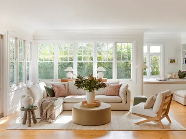 A living room with light timber floorboards, white walls and large windows that look out on to greenery. A cream modular sofa is positioned in the middle, styled with throws and cushions. A round ottoman is positioned in the middle, creating a coffee table that has a cream vase on top, displaying greenery.