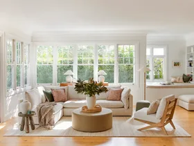 A living room with light timber floorboards, white walls and large windows that look out on to greenery. A cream modular sofa is positioned in the middle, styled with throws and cushions. A round ottoman is positioned in the middle, creating a coffee table that has a cream vase on top, displaying greenery.