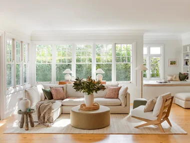 A living room with light timber floorboards, white walls and large windows that look out on to greenery. A cream modular sofa is positioned in the middle, styled with throws and cushions. A round ottoman is positioned in the middle, creating a coffee table that has a cream vase on top, displaying greenery.
