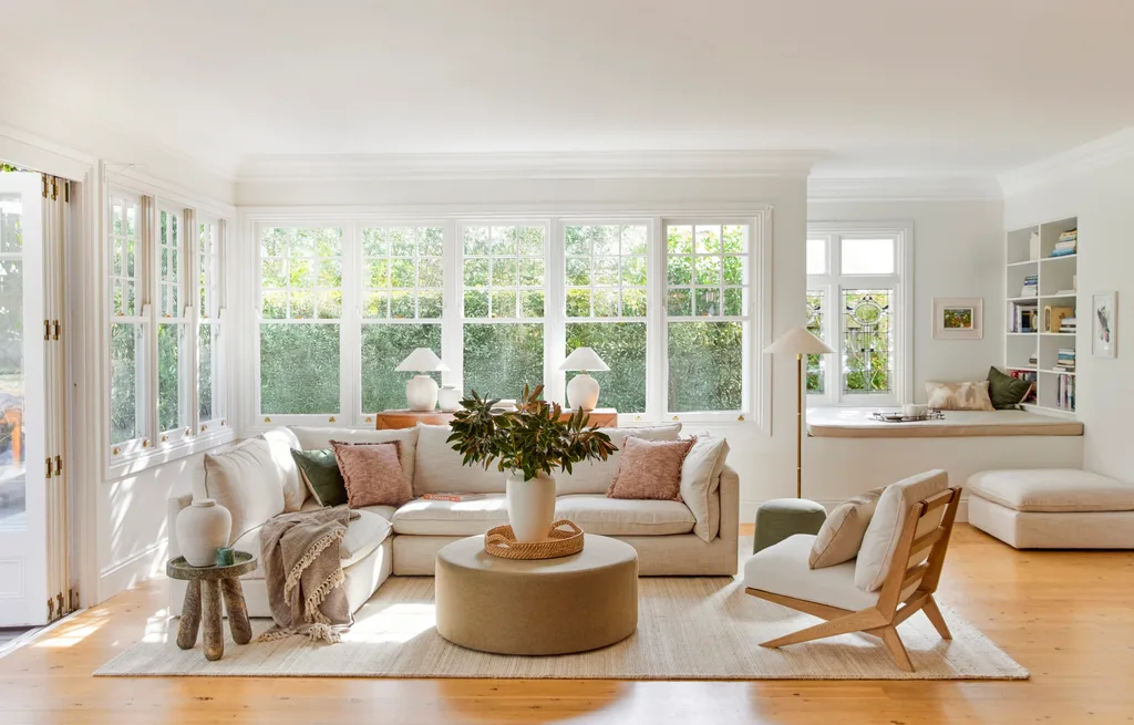 A living room with light timber floorboards, white walls and large windows that look out on to greenery. A cream modular sofa is positioned in the middle, styled with throws and cushions. A round ottoman is positioned in the middle, creating a coffee table that has a cream vase on top, displaying greenery.