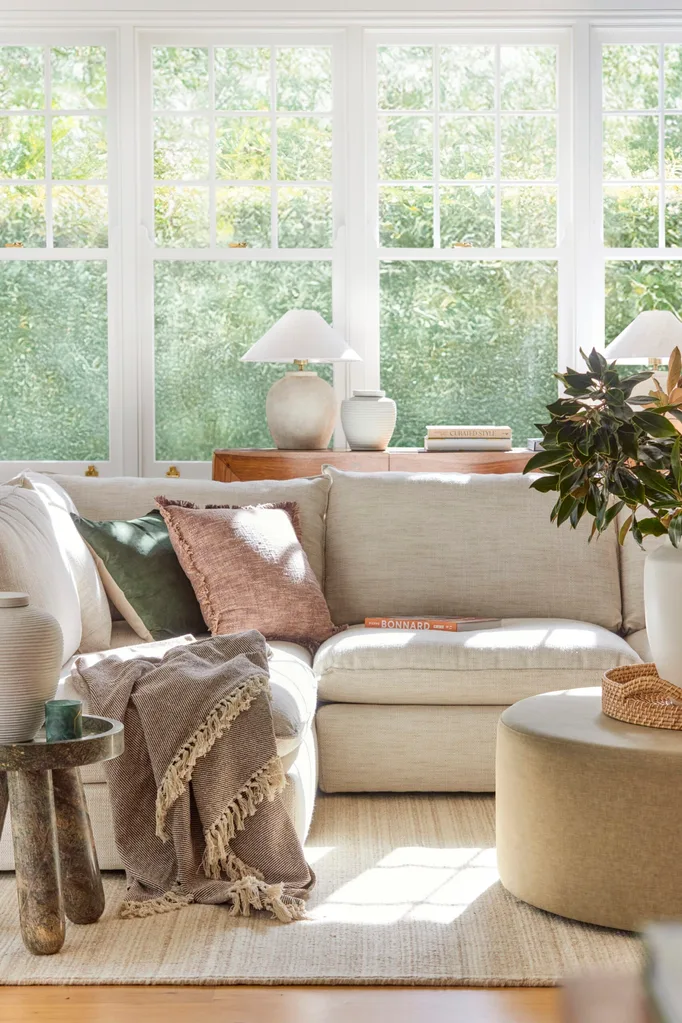 A cream sofa is positioned in front of large windows that look out on to greenery. The couch is styled with throws and cushions. A round ottoman is positioned in the middle, creating a coffee table that has a cream vase on top, displaying greenery.