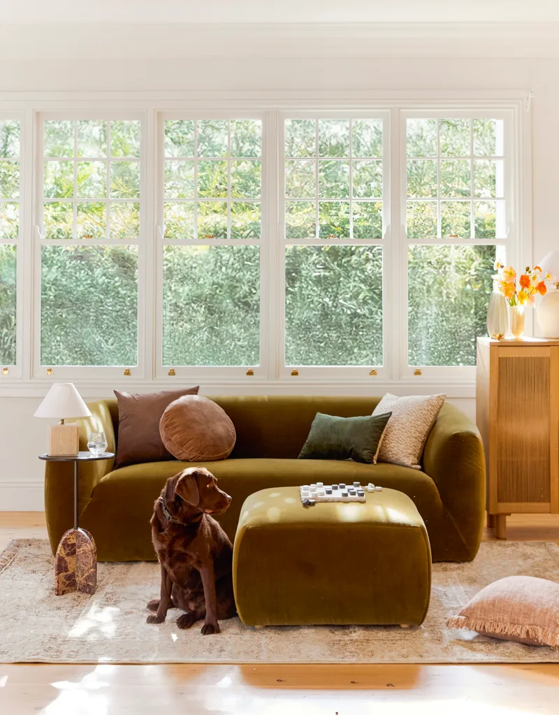 A green sofa that has been styled with a selection of cushions in deep, earthy tones. An ottoman is being used as a coffee table, complete with a board of checkers atop. A dog sits next to the ottoman. This is all positioned in front of a row of windows that look out on to greenery and on top of a traditionally patterned rug.