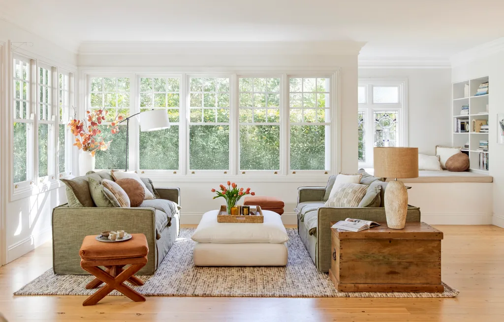 A living room with light timber floorboards, white walls and large windows that look out on to greenery. Two green couches face each other in the middle, styled with throws and cushions. A rectangular ottoman is positioned in the middle, creating a coffee table that has a woven tray positioned on it, with a vase of tulips and candles on it.