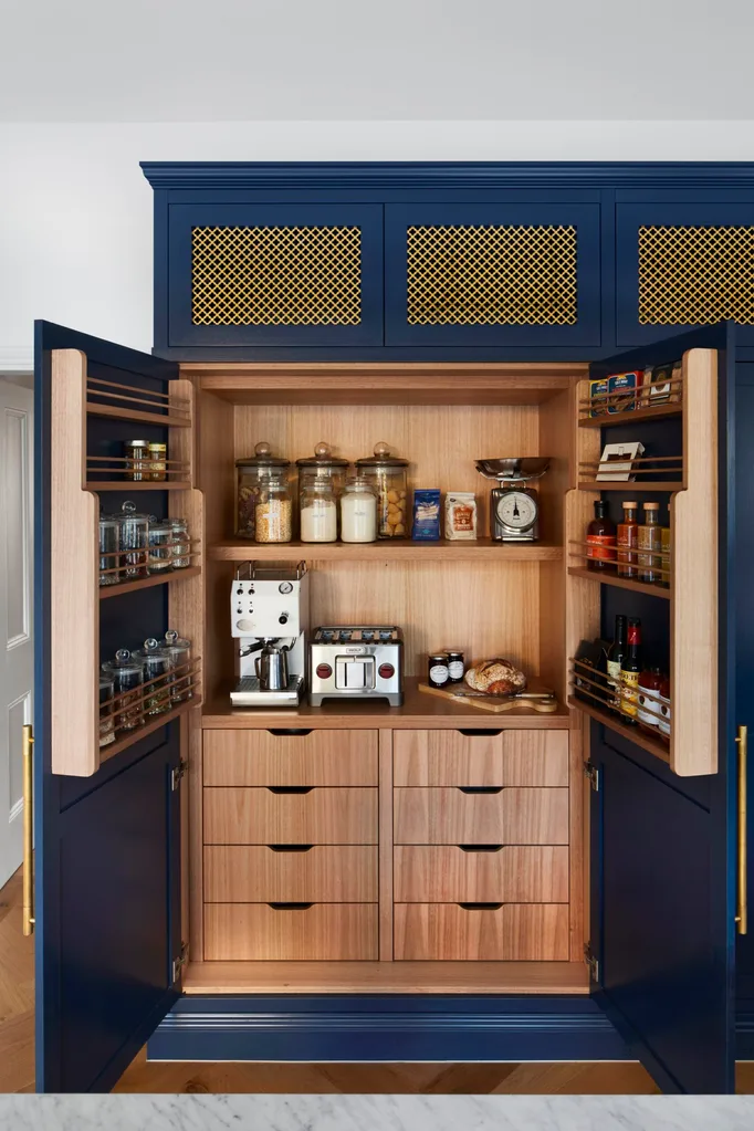 A kitchen with blue coloured joinery and brass mesh profiles. One kitchen cupboard is open to reveal appliances stored inside, such as a coffee machine and toaster. The inside of the joinery is wooden.