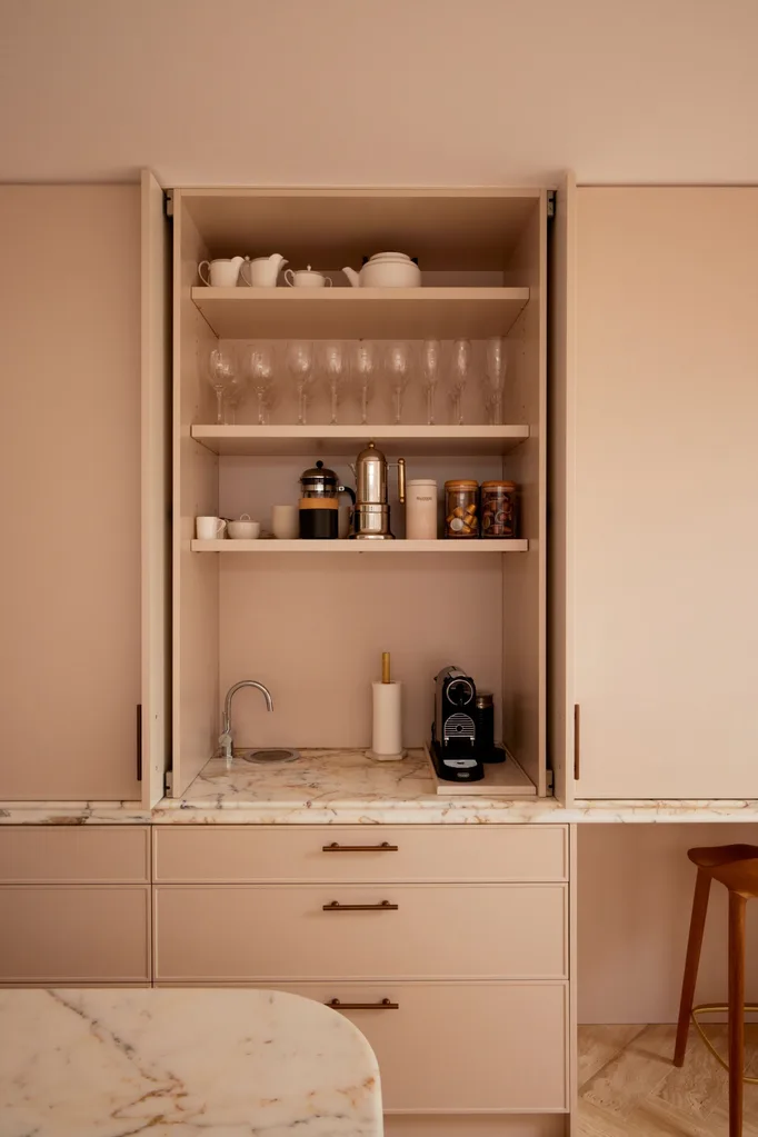 A kitchen with floor to ceiling cupboards. The cabinets are a dusty pink shade with brass pull handles. Two cupboard doors are open to reveal an appliance cupboard. The benchtop is marble, a tap is off to one side and a small coffee machine and accessories are stored in this cupboard.