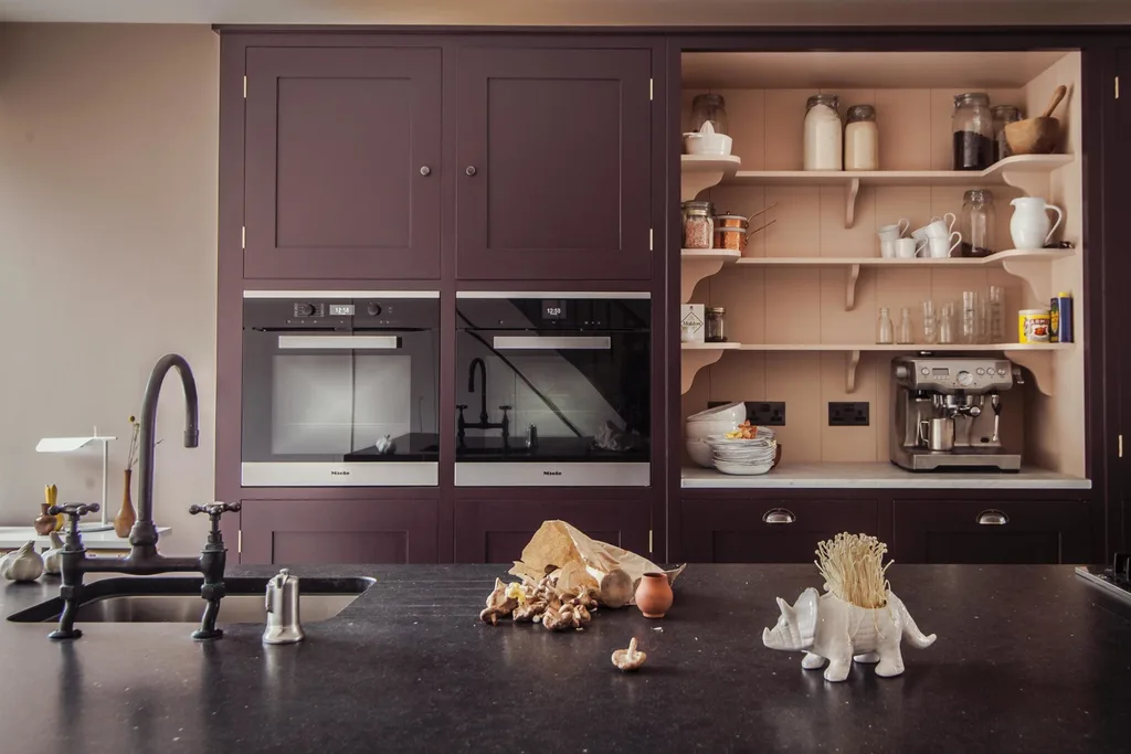 A kitchen with a black island countertop and plum coloured joinery. One side of the kitchen cupboard is open to reveal appliances stored inside, such as a coffee machine and accessories. Power points are inside the kitchen cabinet, which is painted a dusty pink.