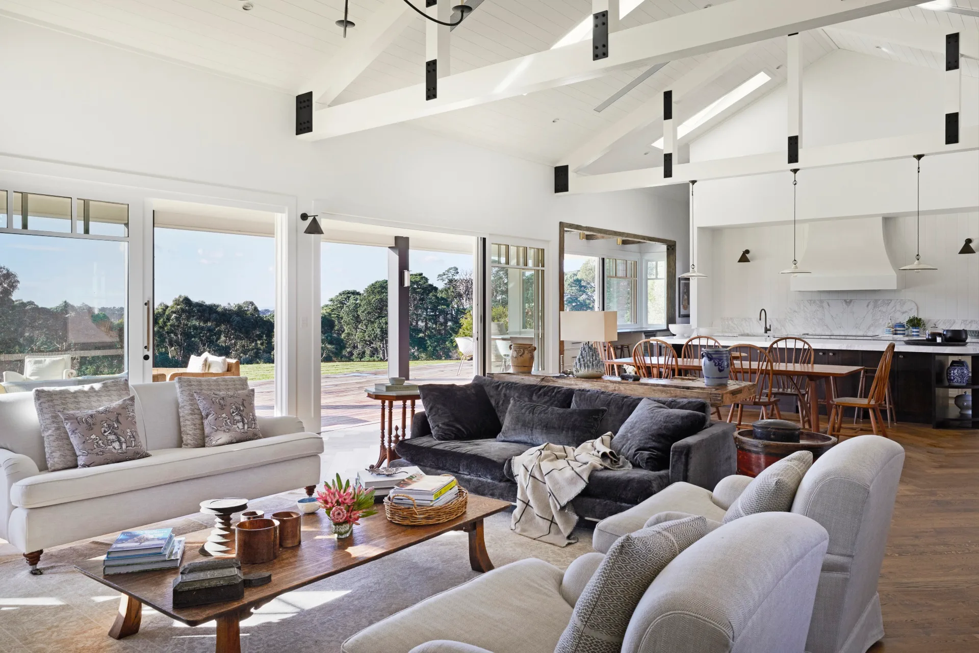 An open-plan living room inside a modern farmhouse with comfortable grey couches and views outside. 