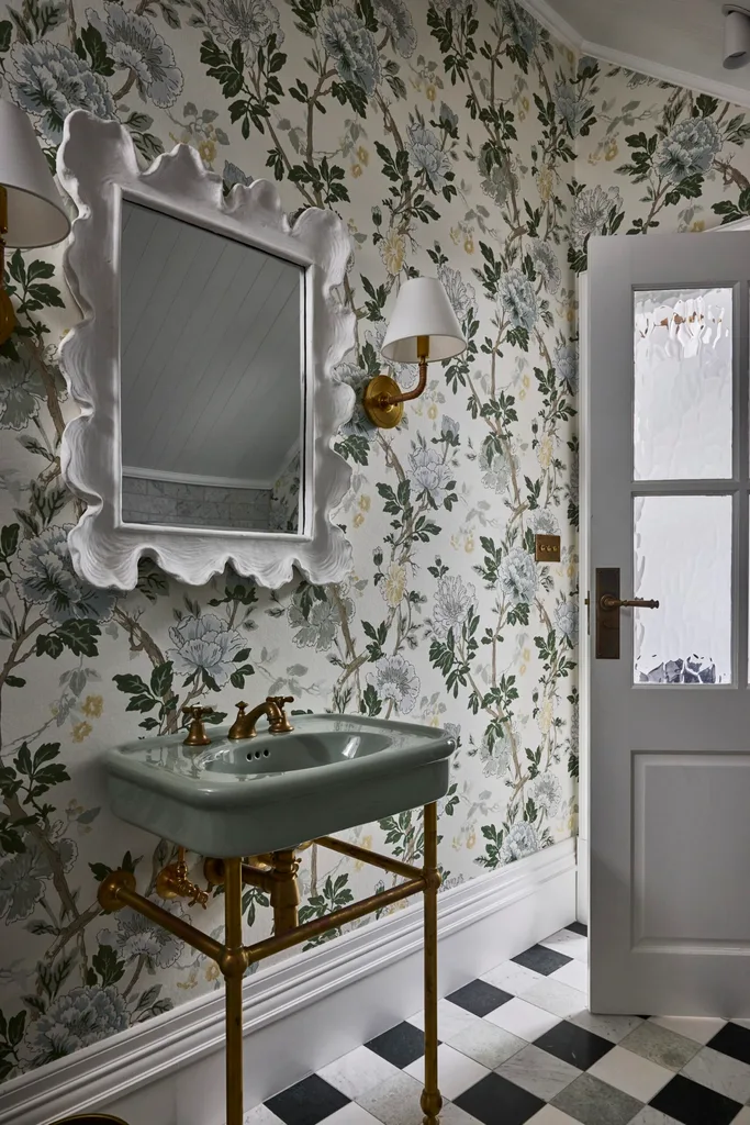 A powder room with checkered floor tiles, a basin on a brass frame and floral wallpaper.