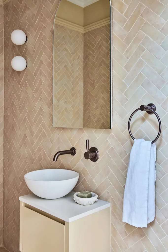 A powder room with sand tone wall tiles (laid in a herringbone pattern), a basin that sits on the benchtop of a vanity and round wall lights beside an arched mirror.
