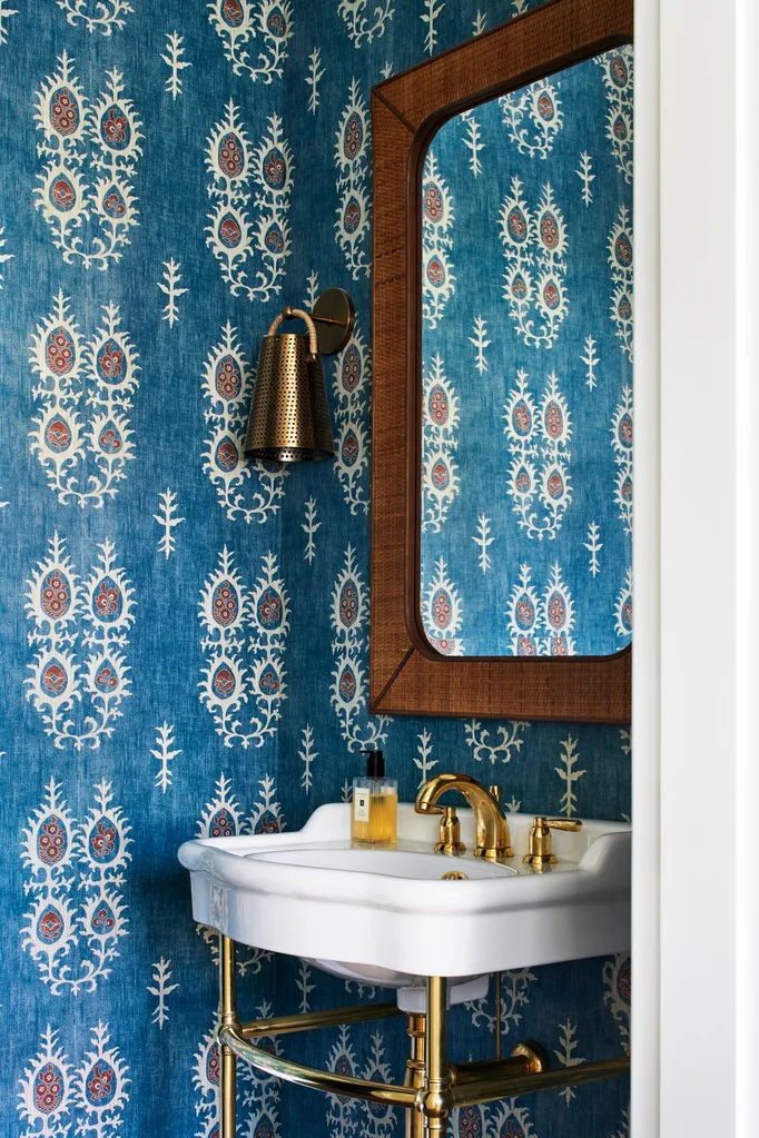A powder room with a basin on a gold frame, blue wallpaper and a rattan mirror frame.