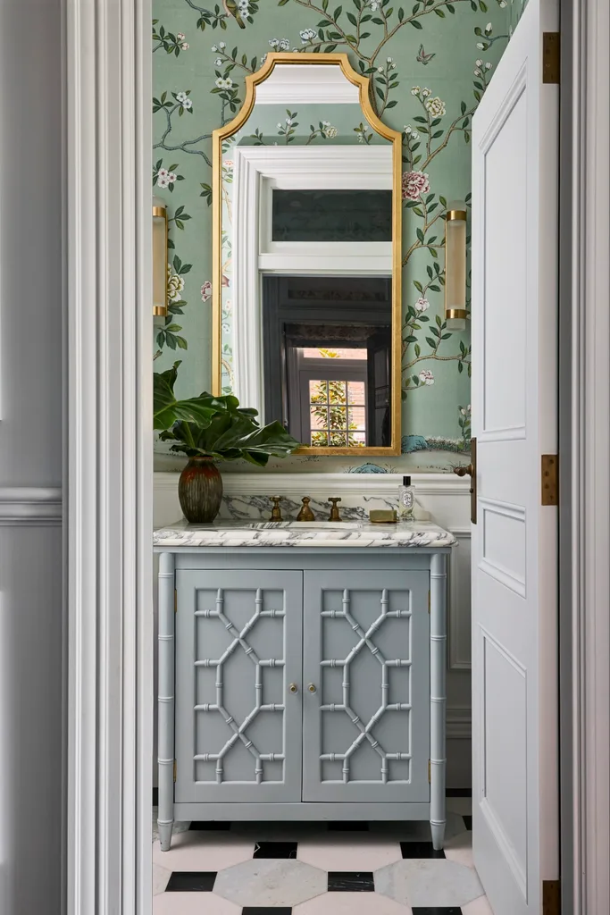 A powder room with wallpaper, wainscoting, a basin built in to a vanity with joinery and a brass-framed mirror.