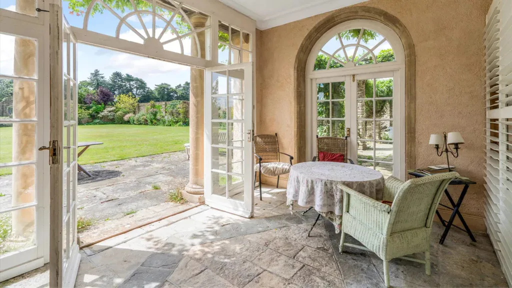 A sunroom with arched French doors that open out to a green garden. A small table with three chairs is positioned in the sunroom, alongside a side table with lamps on top.