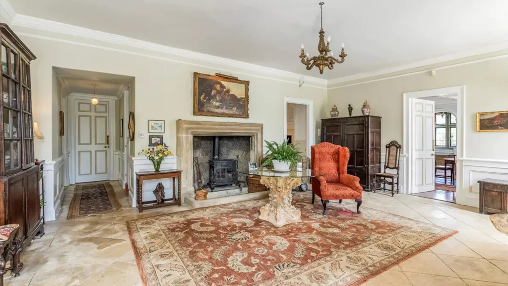 A traditional style sitting area with a grand fireplace, large rug, wingback armchair and oil painting above the fireplace. The walls feature wainscoting with cornices above.