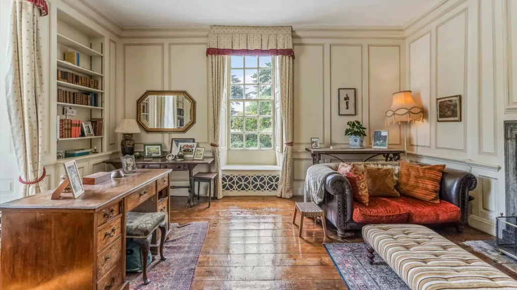 A sitting room with walls that feature decorative moulding, a fireplace, a large wooden desk, a built-in bookshelf and curtains with valances.