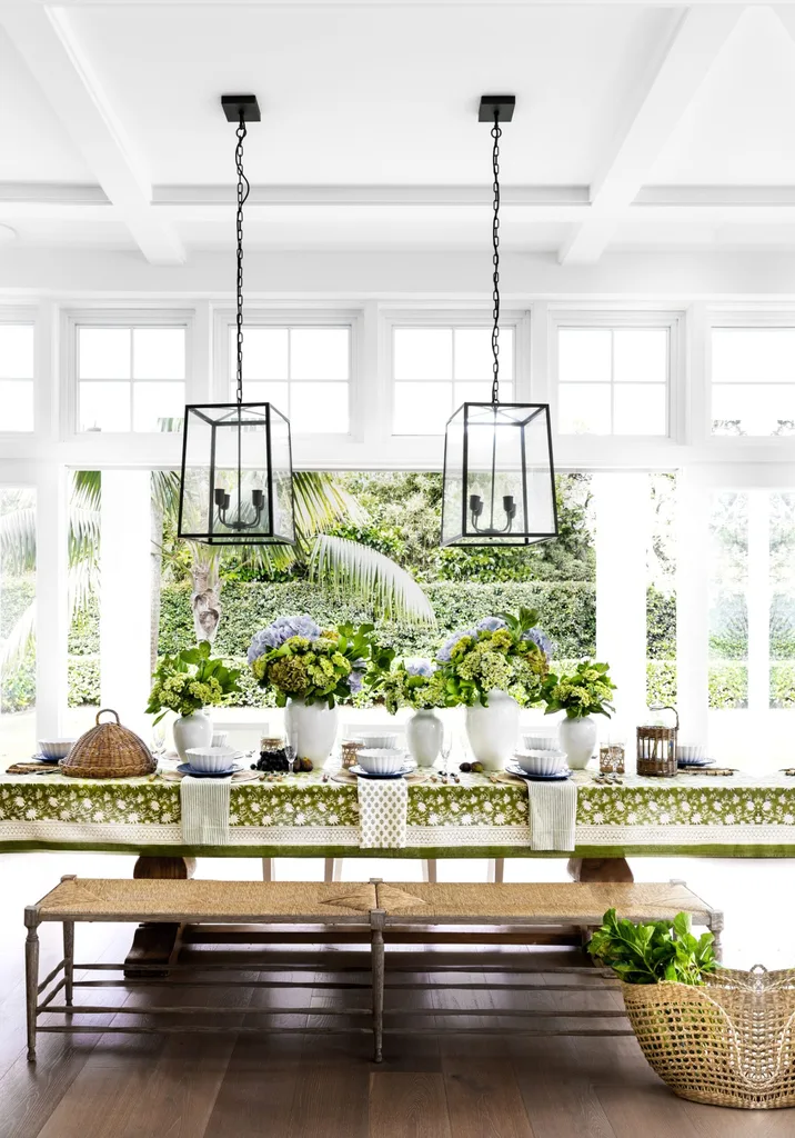 Alfresco dining area. The table is set with floral arrangements, wicker bench, hanging lantern lights, and green patterned tablecloth and clerestory windows above.