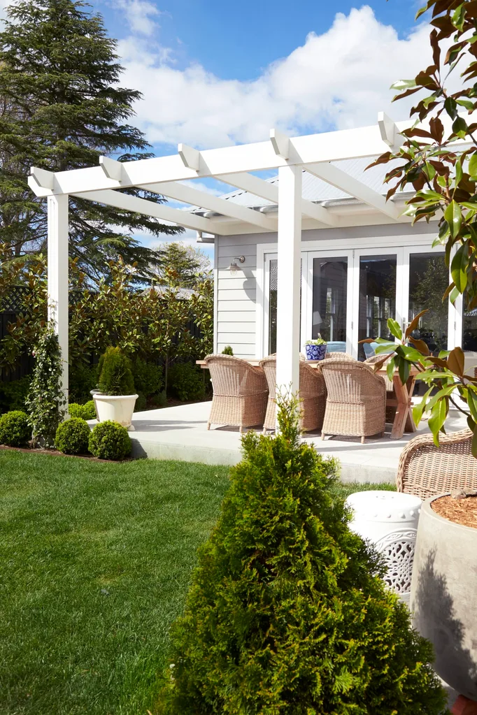 White pergola over an alfresco dining area with wicker chairs, timber dining table, surrounded by lush greenery and a blue sky above with wispy clouds.