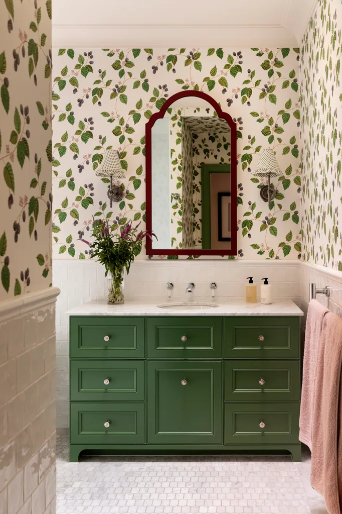 Bathroom with green vanity, floral wallpaper, white tiled wainscoting, red mirror, and a vase of flowers.