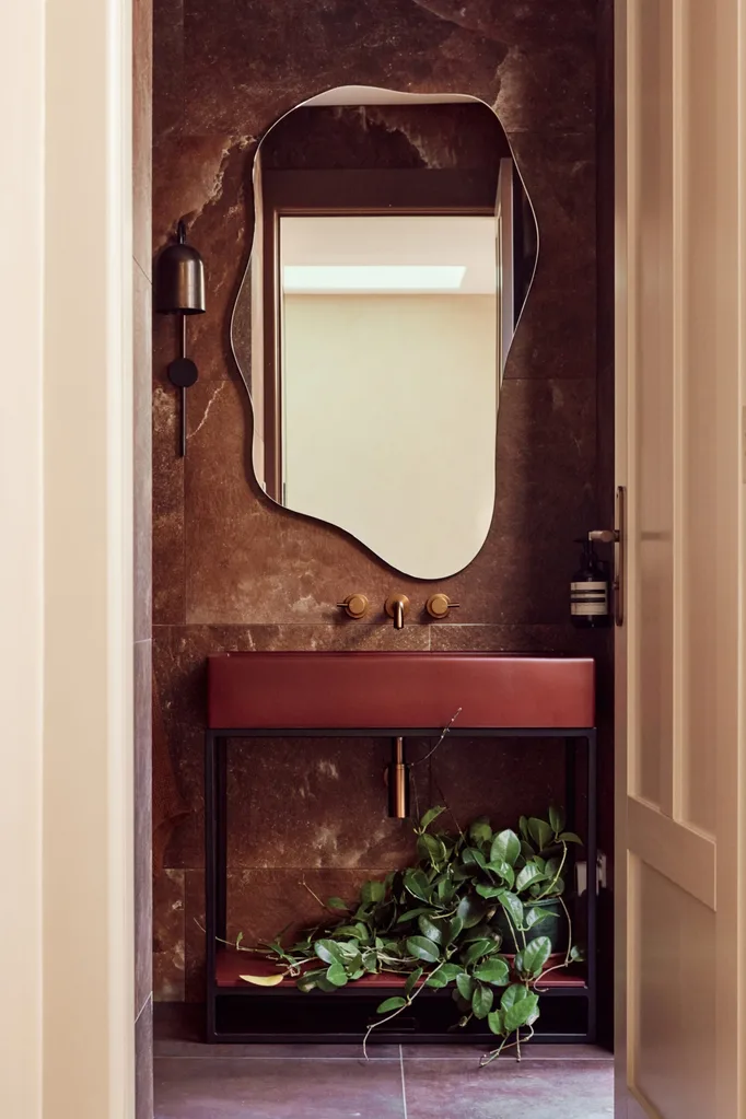 A door is open leading to a powder room with asymmetrical, curved mirror, brown vanity with black frame, one wall sconce, and green plant under the sink.