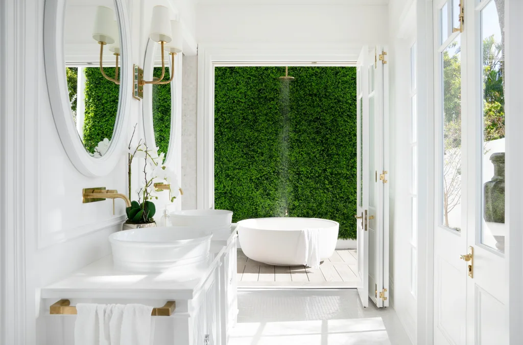 Luxurious white bathroom with double sinks, round mirrors, and brass hardware. The room opens out to a wall of greenery. An outdoor bath, with an outdoor shower positioned above, is in front of this lush wall. There is ample natural light.