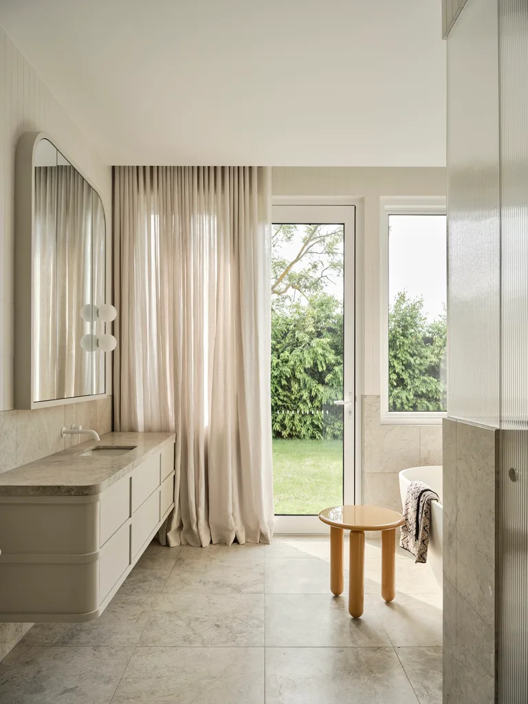 Modern bathroom with a large mirror, beige curtains, floating vanity, round side table positioned next to a bath and a view of a garden. |