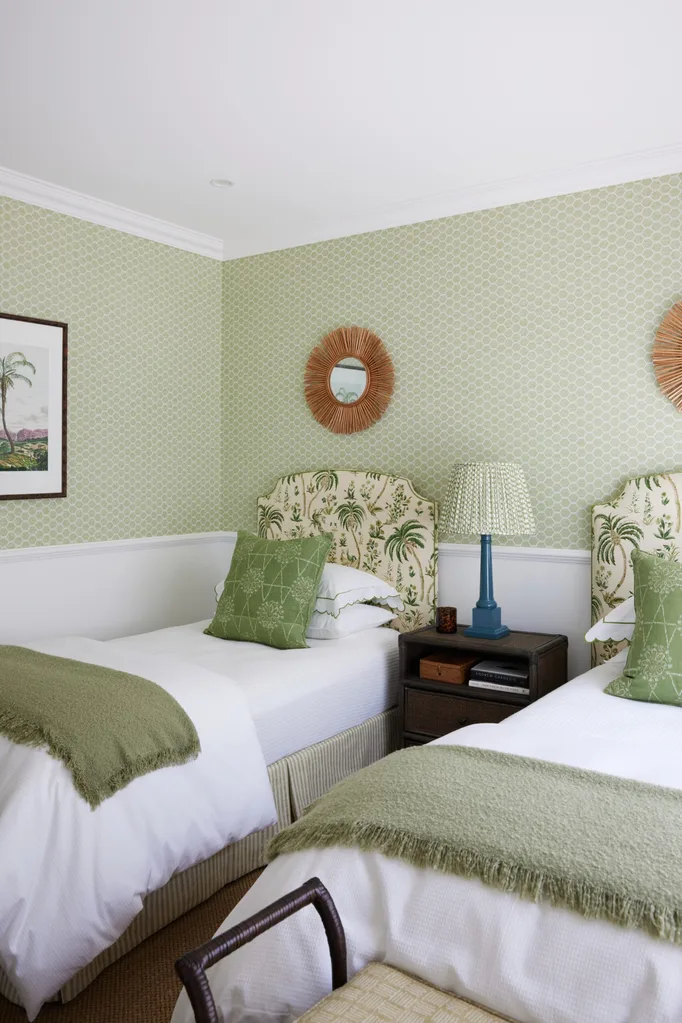 Two twin beds with green patterned wallpaper above white wainscoting, twin rattan mirrors above the twin beds, and a blue lamp on a shared nightstand.