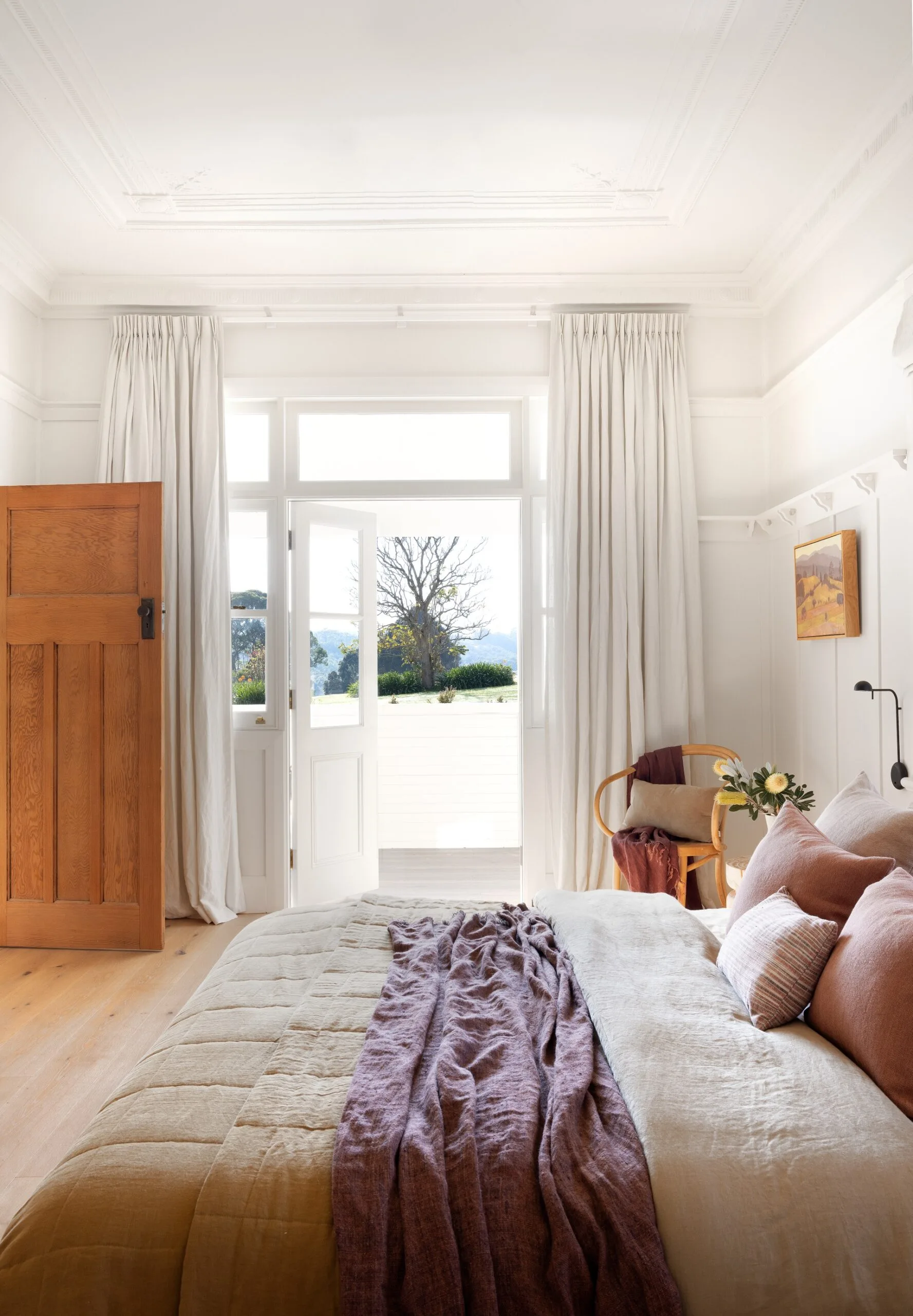 Bright bedroom with white walls, a bed with linen in neutrals and dusty pink, wooden chair in the corner, and large open doors leading to a sunny patio and garden view. |