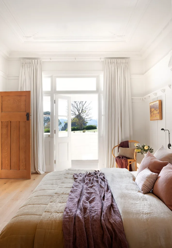 Bright bedroom with white walls, a bed with linen in neutrals and dusty pink, wooden chair in the corner, and large open doors leading to a sunny patio and garden view. |